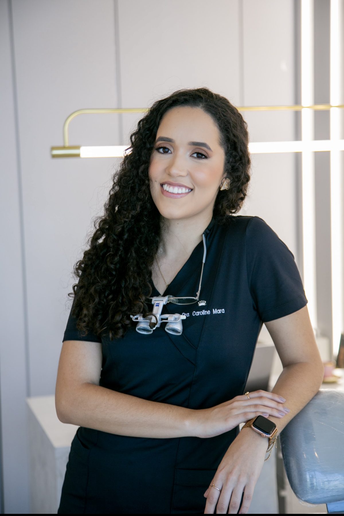 Dra. Caroline Mara com uniforme de atendimento, apoiada na cadeira do consultório